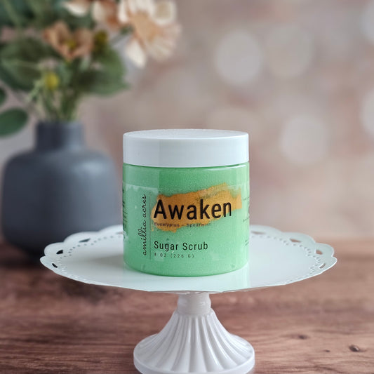 A green jar of Awaken Sugar Scrub by Amillia Acres ~ Goat Milk Soaps and Lotions rests on a white cake stand, ready to reveal glowing skin, with a blurred floral vase and soft, neutral background.
