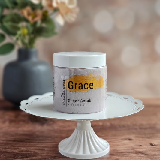 A jar of Grace Sugar Scrub by Amillia Acres ~ Goat Milk Soaps and Lotions rests on a white cake stand, with a blurred gray vase and flowers in the background.