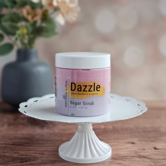 A jar of Dazzle Sugar Scrub by Amillia Acres ~ Goat Milk Soaps and Lotions rests on a white cake stand, with a blurred vase behind it. This exfoliating treat brings a luxurious feel to your skincare routine.