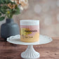 A jar of Amillia Acres ~ Goat Milk Soaps and Lotions "Happiness" Sugar Scrub sits on a white cake stand atop a wooden surface, promising glowing skin with uplifting orange and tangerine scents. Blurred flowers and a vase decorate the background.
