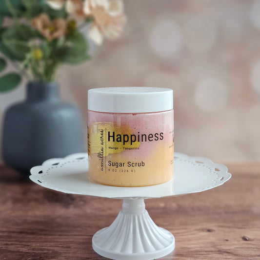 A jar of Amillia Acres ~ Goat Milk Soaps and Lotions "Happiness" Sugar Scrub sits on a white cake stand atop a wooden surface, promising glowing skin with uplifting orange and tangerine scents. Blurred flowers and a vase decorate the background.