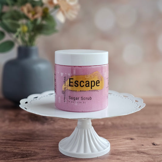 A pink jar of "Escape" Sugar Scrub by Amillia Acres ~ Goat Milk Soaps and Lotions, an exfoliating scrub for glowing skin, sits on a white cake stand with a blurred gray flower vase in the background on a wooden surface.