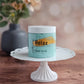 A jar of Amillia Acres ~ Goat Milk Soaps and Lotions "Relax | Sugar Scrub" rests on a white pedestal, with a soft, floral background—showcasing this exfoliating body scrub for glowing skin.