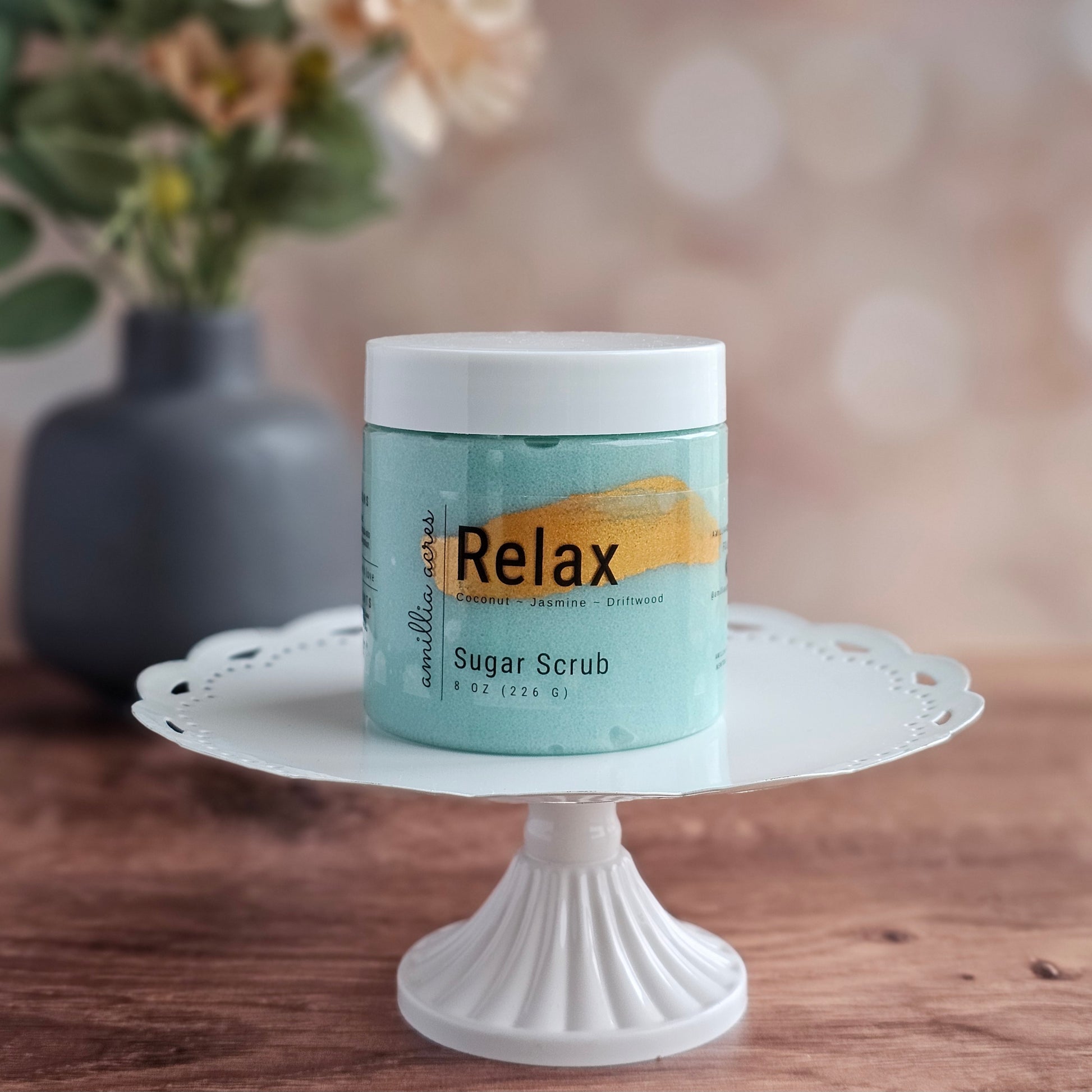 A jar of Amillia Acres ~ Goat Milk Soaps and Lotions "Relax | Sugar Scrub" rests on a white pedestal, with a soft, floral background—showcasing this exfoliating body scrub for glowing skin.