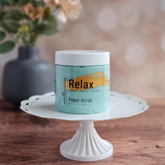 A jar of Amillia Acres ~ Goat Milk Soaps and Lotions "Relax | Sugar Scrub" rests on a white pedestal, with a soft, floral background—showcasing this exfoliating body scrub for glowing skin.
