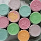 A top view of twelve jars of Amillia Acres ~ Goat Milk Soaps and Lotions' Happiness Sugar Scrub in bright pink, green, blue, and yellow, arranged neatly on a marble surface next to soft green fabric.