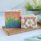 Two handmade soaps sit on a wooden tray. The left one, Flower Child by Amillia Acres, features rainbow swirls and floral decorations. The right soap displays detailed daisy designs with a textured finish. A soft-focus green plant provides a natural backdrop.