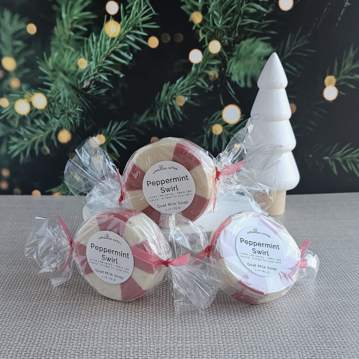 Three round Peppermint Swirl Goat Milk Soaps from Amillia Acres ~ Goat Milk Soaps and Lotions, wrapped in clear plastic with red ribbons, are displayed before a white tree ornament and festive green lights—ideal handmade holiday gifts.