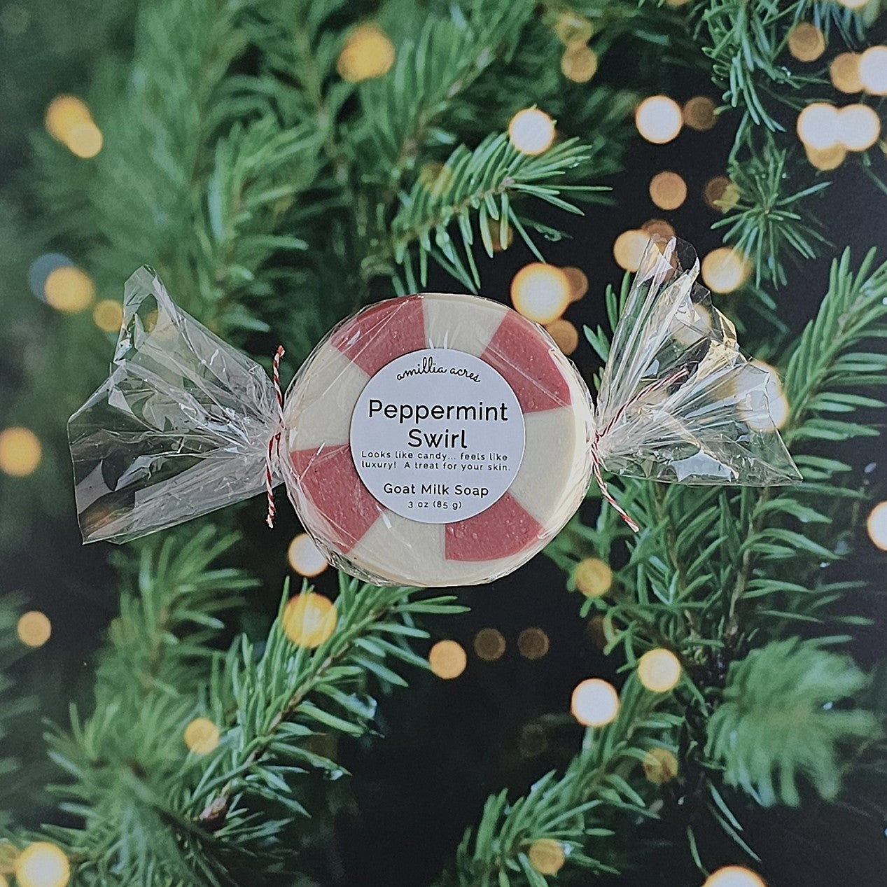 A round Peppermint Swirl Goat Milk Soap by Amillia Acres ~ Goat Milk Soaps and Lotions, wrapped in clear plastic, is displayed before a glowing Christmas tree, its label featuring the festive scent and natural ingredients.