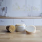A round wooden dish brush and two beige bars of Amillia Acres Solid Dish Soap | Goat Milk Soap sit on a wooden kitchen countertop, with a white ceramic dish and a softly blurred kitchen background behind them.