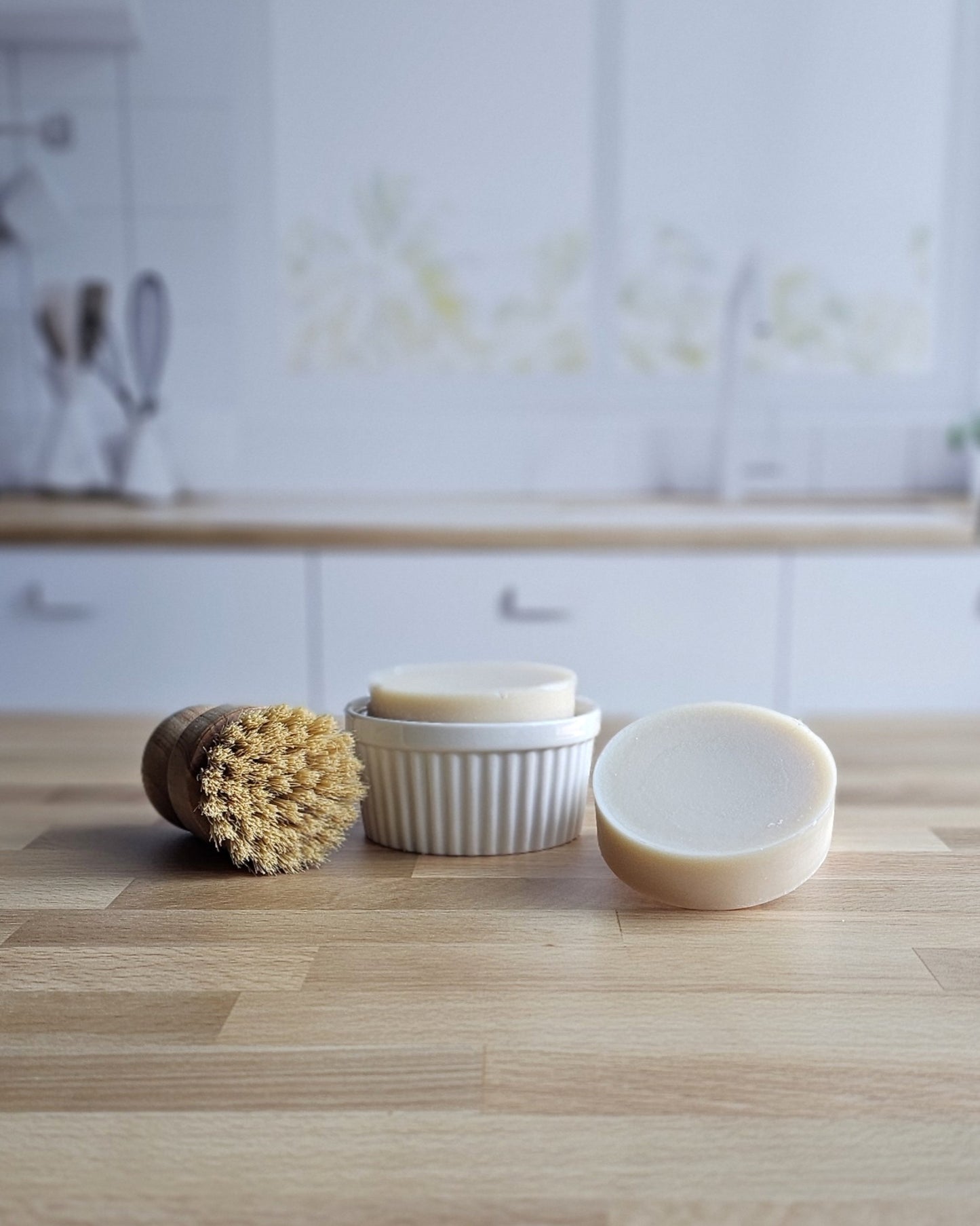 A round wooden dish brush and two beige bars of Amillia Acres Solid Dish Soap | Goat Milk Soap sit on a wooden kitchen countertop, with a white ceramic dish and a softly blurred kitchen background behind them.