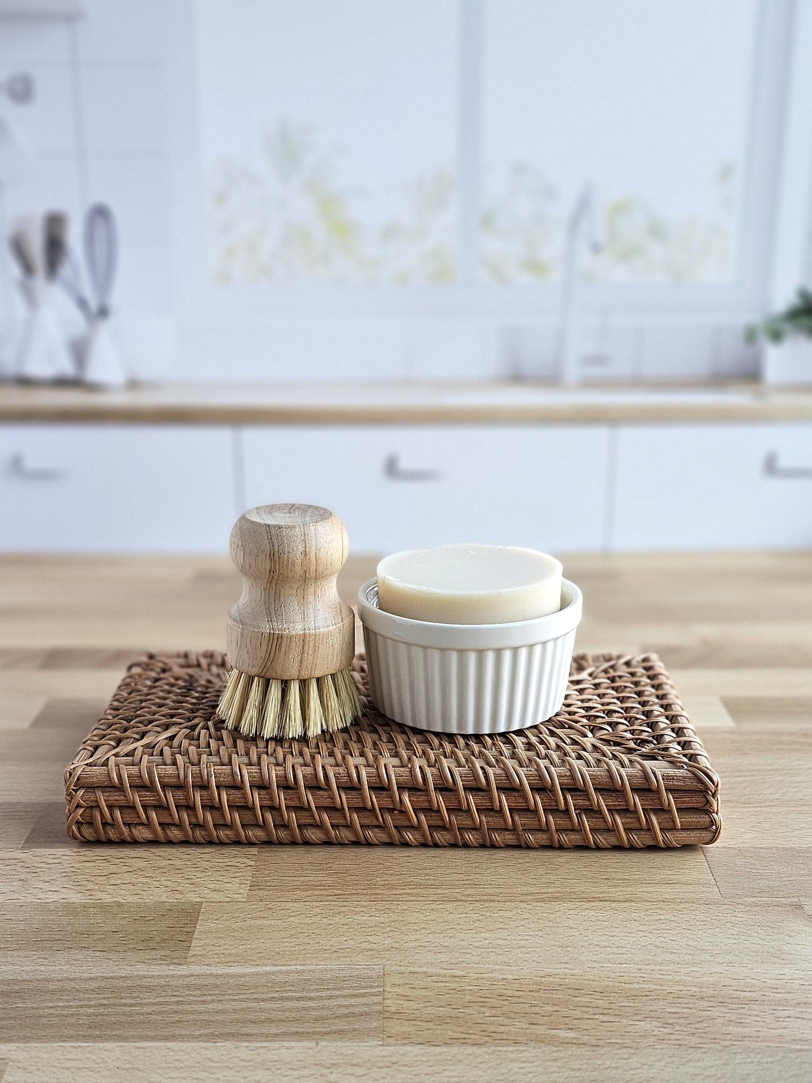 An Amillia Acres Solid Dish Soap bar and a wooden scrub brush rest in a white ramekin on a woven placemat atop a kitchen counter. Brand: Amillia Acres ~ Goat Milk Soaps and Lotions. Product contains nourishing goat milk.