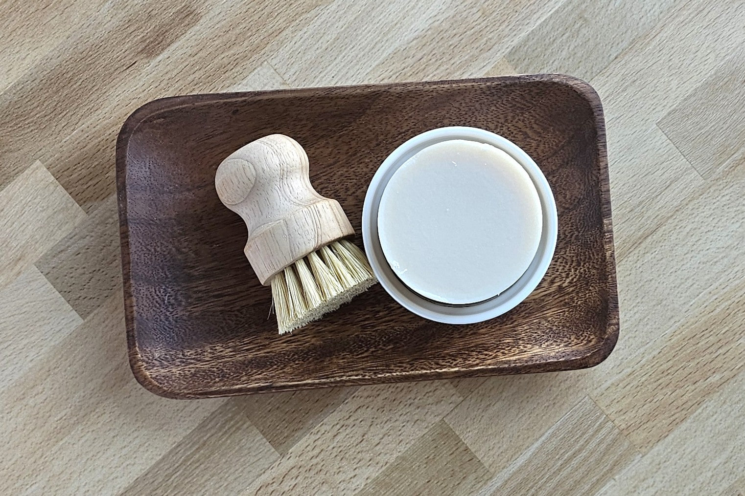 Amillia Acres Solid Dish Soap, a powerful grease-cutting goat milk soap, rests in a white dish next to a wooden scrub brush on a dark tray over light wood.