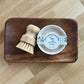 A wooden tray holds a 3.5 oz Solid Dish Soap in a white tin labeled "Amillia Acres ~ Goat Milk Soaps and Lotions," next to a small wood scrub brush with light bristles—perfect for grease-cutting cleaning.