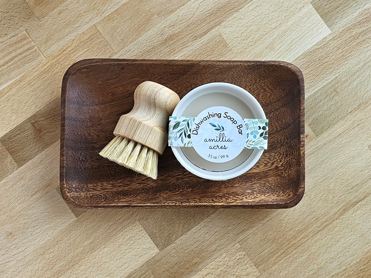 A wooden tray holds a 3.5 oz Solid Dish Soap in a white tin labeled "Amillia Acres ~ Goat Milk Soaps and Lotions," next to a small wood scrub brush with light bristles—perfect for grease-cutting cleaning.