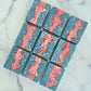 Nine rectangular Liberty bars from Amillia Acres feature blue, speckled white, and abstract pink swirls. Arranged on marble, these handmade goat milk soaps showcase artisanal craftsmanship and natural beauty.