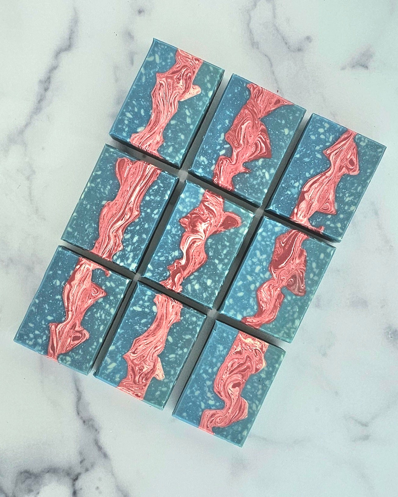 Nine rectangular Liberty bars from Amillia Acres feature blue, speckled white, and abstract pink swirls. Arranged on marble, these handmade goat milk soaps showcase artisanal craftsmanship and natural beauty.