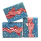 Three rectangular bars of Liberty Goat Milk Soap by Amillia Acres, featuring a blue base, white speckles, and a swirling red-pink marbled center, are stacked against a white background.