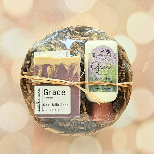 The Small Gift Basket by Amillia Acres ~ Goat Milk Soaps and Lotions features a "Grace" lavender goat milk bar soap, matching body lotion, exfoliating soap saver pouch, brown shredded paper in a basket, and raffia tie.