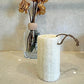 The Amillia Acres Natural Loofah Sponge, a cylindrical eco-friendly scrubber with a rope loop, rests on a countertop. Behind it, a clear glass vase holds dried brown flowers against a neutral wall.