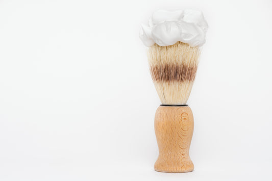 The Amillia Acres Classic Shave Brush features a wooden handle and upright bristles crowned with rich white shaving cream, displayed against a plain white background—a grooming essential.
