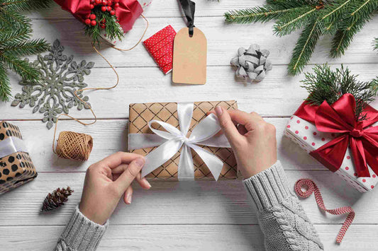 Hands tying a white ribbon on an Amillia Acres Custom Gift Box, specially wrapped in plaid as part of their Custom Gift Box Service Add-On for personalized orders. The scene is adorned with festive items like a snowflake ornament, pinecones, small gift boxes, and greenery. A blank gift tag and spool of twine lie on the white wooden surface, highlighting the unique charm of personalized gifting.