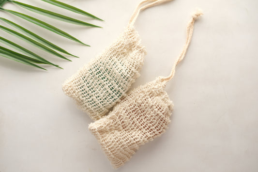 Two Amillia Acres Exfoliating Soap Saver Bags in natural woven mesh with drawstrings are displayed on a light surface, with a green palm leaf visible in the top left corner.
