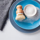 The Amillia Acres Dish Scrubber with durable bristles rests on a blue plate next to a bowl of soapy foam and bubbles, accompanied by a gray textured cloth on a white surface.