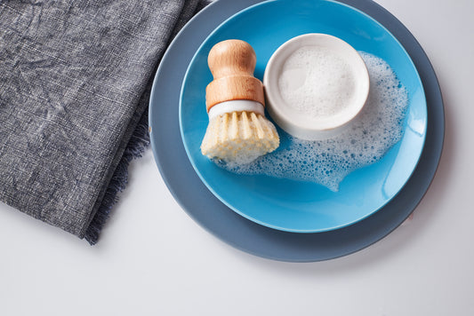 The Amillia Acres Dish Scrubber with durable bristles rests on a blue plate next to a bowl of soapy foam and bubbles, accompanied by a gray textured cloth on a white surface.
