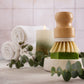 The Amillia Acres Dish Scrubber rests on a green soap bar in a dish, with eucalyptus leaves, rolled white towels, and a lit candle in the background on a light surface.