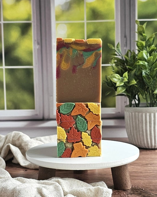 Two handmade Autumn Goat Milk Soaps from Amillia Acres are stacked on a white round stand, each adorned with colorful leaf designs. A potted plant and a sunny green view appear in the background.