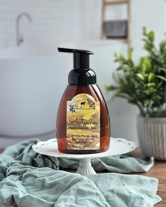 An Amillia Acres ~ Goat Milk Soaps and Lotions "Autumn" Foaming Hand Soap rests on a white pedestal beside a teal towel, with a potted plant in the background.