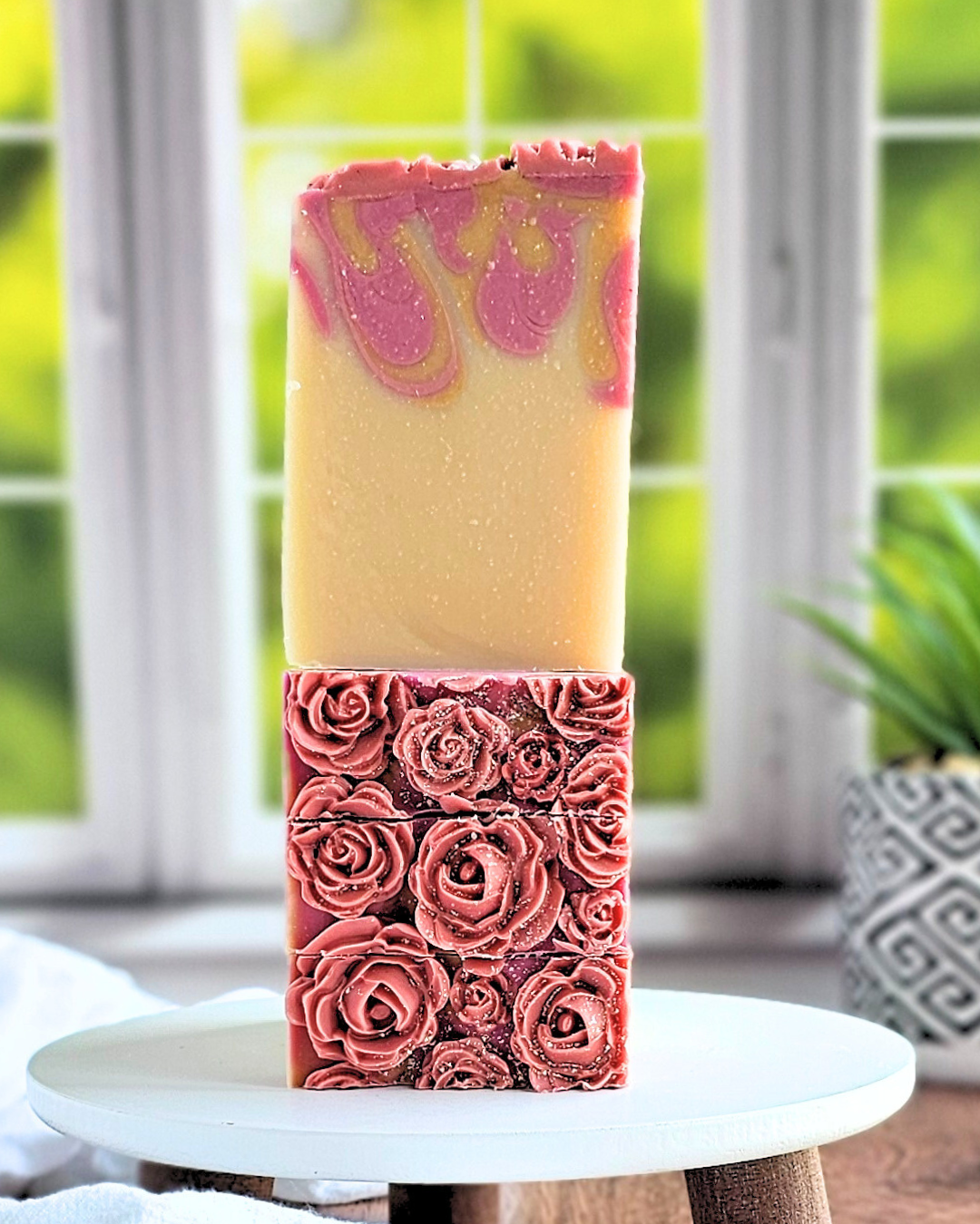 Two Blossom | Goat Milk Soap bars from Amillia Acres are stacked vertically: the top features a handmade yellow bar with pink swirls crafted from natural oils, while the bottom has intricate pink rose designs. They sit elegantly on a white circular stand against a blurred window and potted plant backdrop.