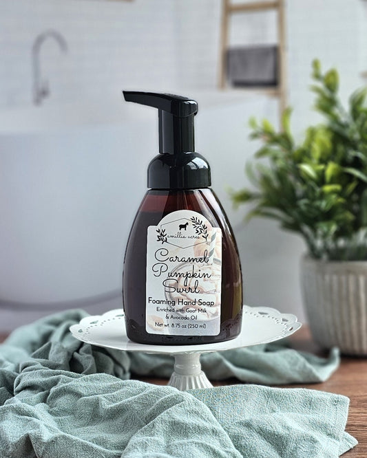 A bottle of Amillia Acres ~ Goat Milk Soaps and Lotions Caramel Pumpkin Swirl Foaming Hand Soap sits on a white cake stand with a green cloth nearby, in a bathroom setting, highlighting this moisturizing hand soap.