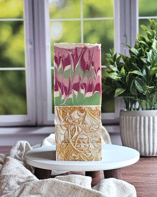 Three Enchanted Goat Milk Soap bars by Amillia Acres ~ Goat Milk Soaps and Lotions are stacked on a white stand; the top bar shows pink, green, and white swirls, while the two below have gold details. A green plant and window sit in the background.