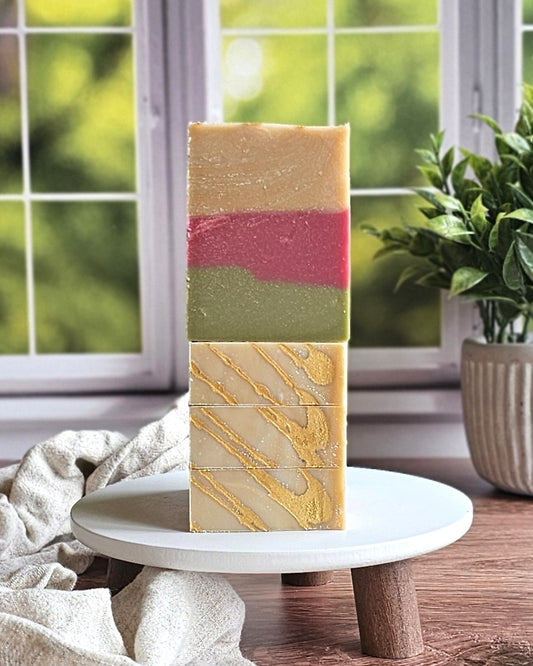 Three Farmstand Apple goat milk soaps by Amillia Acres sit stacked on a white round stand. The artisanal cream bars with gold drizzle and a touch of color are set beside a plant and window, adding a fresh, natural vibe.