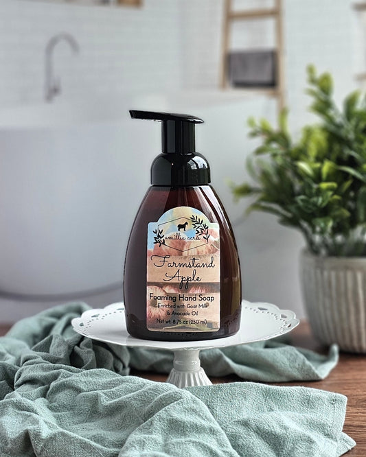 A bottle of Amillia Acres ~ Goat Milk Soaps and Lotions Farmstand Apple Foaming Hand Soap sits on a white cake stand, with a green cloth beside it and a softly blurred bathroom with plants in the background.