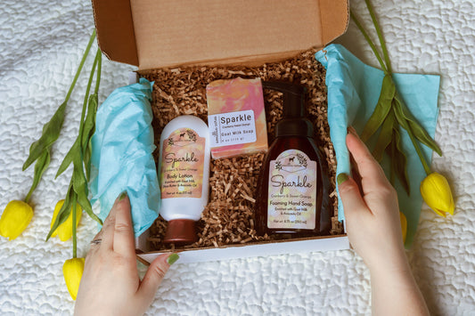 Someone opens the Amillia Acres ~ Goat Milk Soaps and Lotions Signature Gift Box Set (3 piece) with scented soap, body lotion, and foaming hand soap amid blue tissue paper, yellow tulips, and brown crinkle paper on a white textured surface.