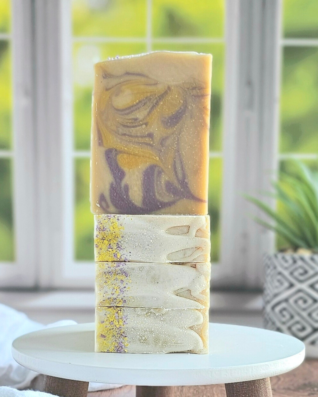 Three Amillia Acres ~ Goat Milk Soaps in earthy tones and swirled patterns are stacked by a window. The top Meadow soap showcases purple and gold swirls, with greenery and a patterned pot in the blurry background.