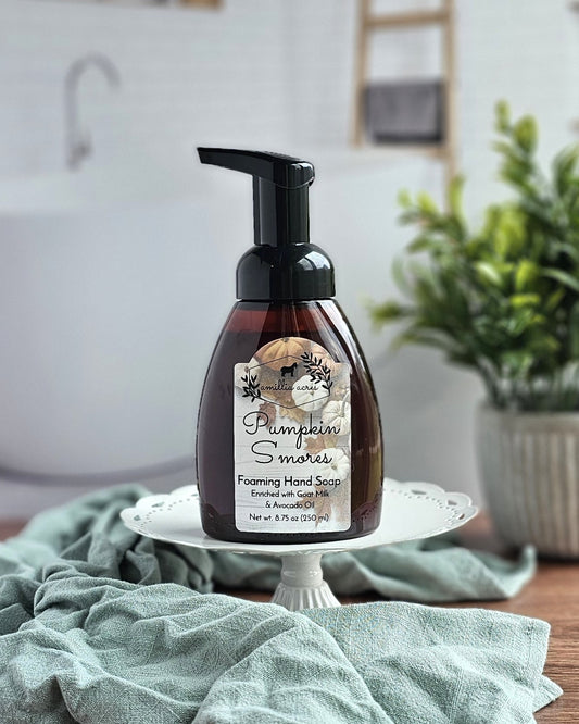 A bottle of Amillia Acres ~ Goat Milk Soaps and Lotions Pumpkin S'mores Foaming Hand Soap rests on a white pedestal by a green towel, ready to provide a gentle, moisturizing cleanse with every wash.