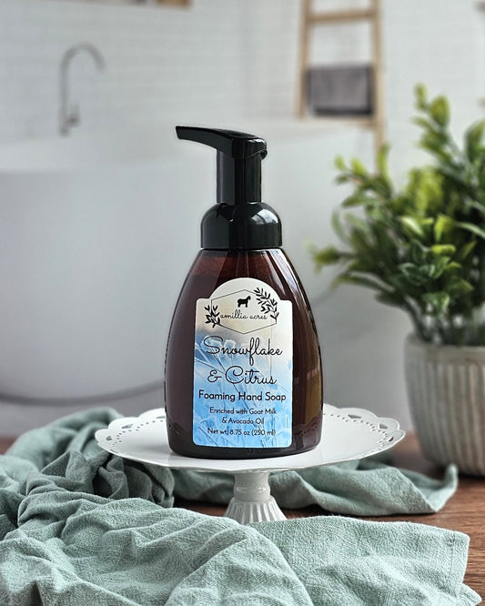 A brown bottle of Amillia Acres Snowflake & Citrus Foaming Hand Soap rests on a white stand in a bathroom, with a potted plant beside it and a blurry bathtub in the background.