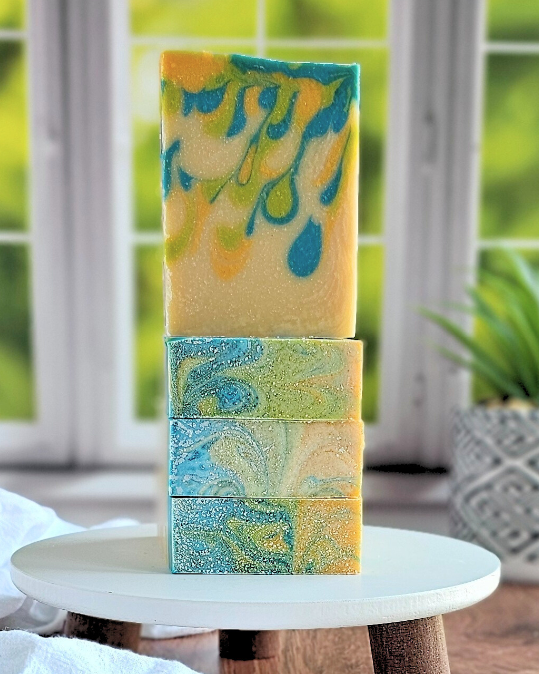 A collection of Tropics Goat Milk Soaps from Amillia Acres sits on a small round white platform, showcasing swirling designs in blue, green, and yellow. Made with natural oils, these artisanal bars are framed by a blurred window and greenery, creating a soothing backdrop.