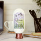 A bottle of Amillia Acres Grace Goat Milk Lotion sits on a desk alongside a laptop, mug, utensil holder, and notebook. Its label features goat milk, shea butter, and avocado oil for a soothing handmade moisturizer.