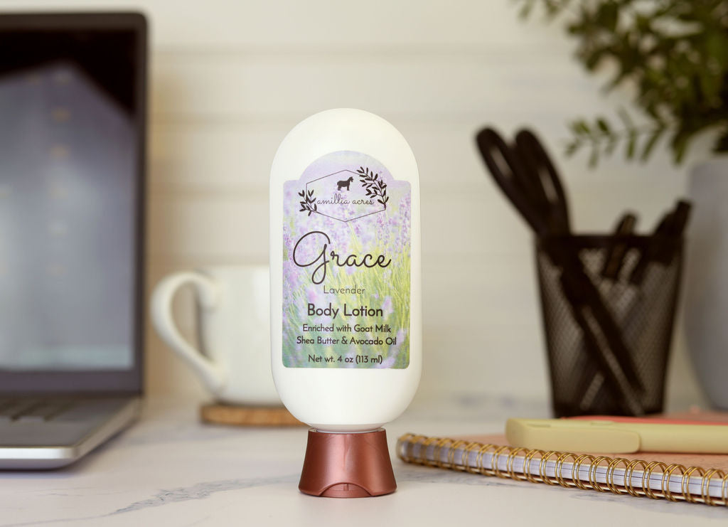A bottle of Amillia Acres Grace Goat Milk Lotion sits on a desk alongside a laptop, mug, utensil holder, and notebook. Its label features goat milk, shea butter, and avocado oil for a soothing handmade moisturizer.