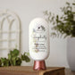 A white bottle of Amillia Acres Simplicity Goat Milk Lotion with a copper cap rests on stacked books. The label features nourishing goat milk, shea butter, and avocado oil. Blurred green plants appear in the background.