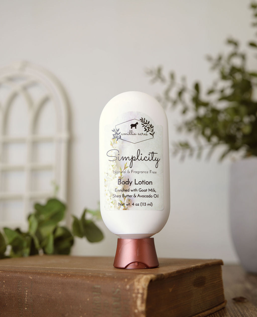 A white bottle of Amillia Acres Simplicity Goat Milk Lotion with a copper cap rests on stacked books. The label features nourishing goat milk, shea butter, and avocado oil. Blurred green plants appear in the background.