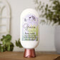 A bottle of Amillia Acres Grace Goat Milk Lotion with a purple and green label sits on a brown surface, against a softly blurred background featuring greenery and a white window.