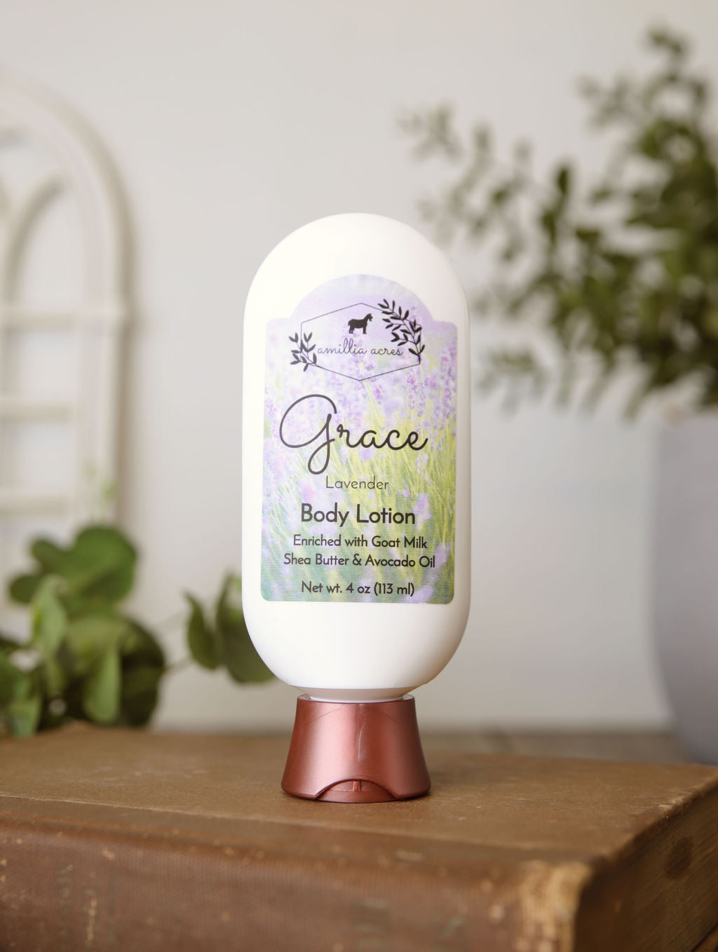 A bottle of Amillia Acres Grace Goat Milk Lotion with a purple and green label sits on a brown surface, against a softly blurred background featuring greenery and a white window.