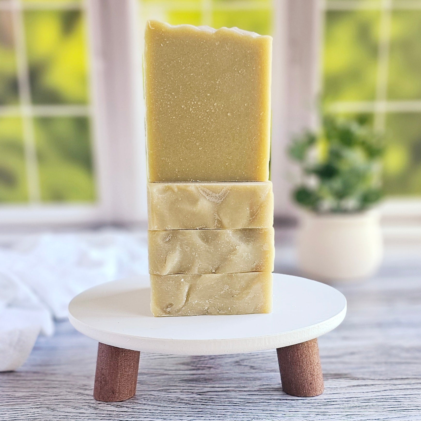 Three bars of Avocado & Yogurt Goat Milk Soap from Amillia Acres ~ Goat Milk Soaps and Lotions are elegantly stacked on a small round wooden stand with short legs. The background showcases a blurred view of a window and a potted plant, highlighting a fresh, natural ambiance enriched with the purity of natural oils.