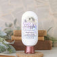 A bottle of Amillia Acres Dazzle Goat Milk Lotion sits on a round wooden stand, with blurred books and green leaves in the background. The label features shea butter, avocado oil, and highlights this rich handmade moisturizer.